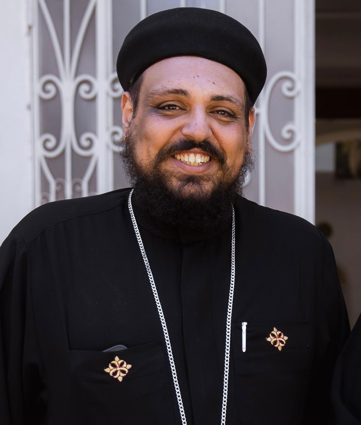 A person wearing a black robe and hat, with a large cross necklace, standing in front of a decorative metal gate.